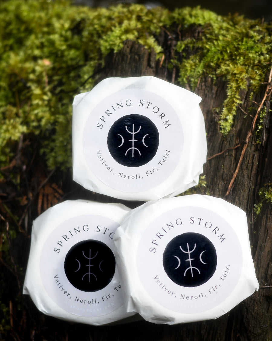 Spring Storm bath bomb with Vetiver, Neroli, Fir, Tulsi by O Apothecary resting by a creek on a log